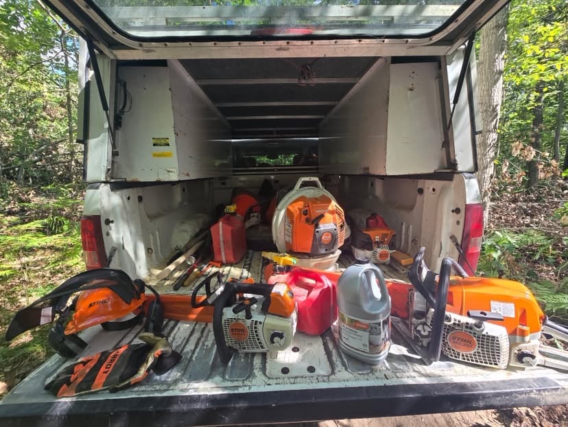Open white trailer loaded with orange and red chainsaws, gas cans, and forestry equipment parked outdoors